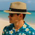 A straw Panama beach hat in tan with a black band, worn by a man in sunglasses and a floral shirt.