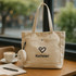 A beige canvas tote bag with two handles and a logo, alongside a cup of coffee and a notebook.
