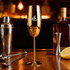 A golden stainless steel champagne flute with a shiny finish, surrounded by bar tools and a bottle of liquor.