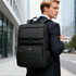 A black laptop backpack is worn by a man in a suit, standing in an urban setting. The backpack features a logo and multiple compartments.