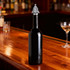 A drink bottle with a silver Christmas tree-shaped wine stopper on a wooden bar top.