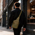 A tactical sling bag in olive green is worn across the back of a person walking on a city street.