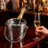 A stainless steel cooler bucket filled with ice, holding a bottle of champagne, beside a champagne flute.