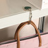 A heart-shaped, silver foldable desk hook is attached to a shelf, holding a pink handbag with brown handles.