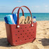 A maroon beach bag with white handles, containing a blue and white towel, flip-flops, and a drink bottle on the sand by the sea.