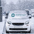 A white car covered with a silver aluminium snow cover, set against a snowy urban backdrop. The cover has a logo.