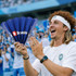 A person with curly hair holds a blue Clap Banner, smiling enthusiastically amidst a cheering crowd.