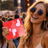 A woman wearing sunglasses holds a red custom-shaped paper fan featuring heart designs, set against a festival backdrop.