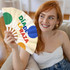 A hand fan featuring colourful circles in green, orange, and blue, held by a smiling person with red hair.