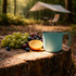A stainless steel insulated mug in teal sits beside a mix of grapes and an orange on a wooden stump in a forest setting.