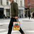 A person walking on the street carries a small, colourful tote bag featuring illustrated characters and a logo.