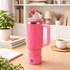 A pink silicone boot for a tumbler, featuring a handle and a lid, set on a wooden table with decorative items.