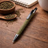 A green pen with a silver tip rests on a wooden surface, surrounded by tea grounds and a notebook.