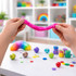 A pair of hands stretches bright pink putty, surrounded by colourful decorative items and a rainbow toy on a table.