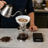 A stainless steel coffee filter with stand is shown, with a kettle pouring water over it. Coffee beans are scattered nearby.