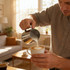 A stainless steel milk frothing pitcher being used to pour milk into a coffee cup in a warm, sunlit living room.