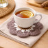 A tea cup with lemon sits on a round, mauve crochet coaster, next to a beige napkin on a wooden table.