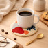 A white coffee cup sits on a multi-coloured punch needle coaster, with a cookie and a spoon nearby on a wooden surface.