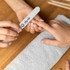 A pack of 50 wooden straight shape nail files in white, resting on a grey towel, with hands in use.
