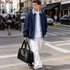 A person in a navy jacket and white pants carries a black leather carry-on duffle bag while walking on the street.