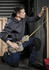 A man in a grey hoodie and dark trousers measures a wooden plank while kneeling, wearing a tool belt.