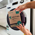 Eco-friendly laundry detergent strips in brown packaging with a logo, held in hands near a washing machine.