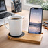 A white mug sits on a bamboo cup warmer with a phone charging beside it. The setting includes a laptop and office supplies.