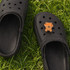 A pair of black clog shoes featuring a 3D bear charm on one shoe, set on green grass.