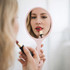 A pink handheld makeup mirror reflects a person applying lipstick, with a soft, neutral background.