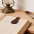 A brown PU leather magnetic bookmark rests on an open book, with a vintage lamp and feather nearby.