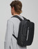 A black backpack with multiple compartments is worn on the back of a person in a grey top. The backpack features a logo.