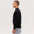 A black crewneck sweatshirt is worn by a model standing sideways, paired with grey sweatpants. The sweatshirt has a logo.
