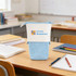 An expandable pencil case in light blue with a white band and a logo, set on a classroom desk with books and stationery.