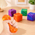 A hand squeezes an orange, marbled cube amidst vibrant purple, green, and blue cubes on a wooden surface.