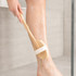 A detachable shower brush with a long wooden handle and white bristles, being used on a leg in a shower setting.
