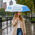 A light blue and white folding umbrella held by a woman wearing a beige trench coat, standing in the rain.