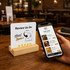 A NFC acrylic menu in white sits on a wooden stand beside a hand holding a smartphone displaying a food ordering app.