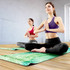 A yoga mat in green with floral patterns, used by two women seated in a meditative pose on a wooden floor.