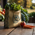 A jute tote bag with a logo, filled with leafy greens, a red apple, and two carrots, set on a wooden table.