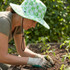 A wide-brim hat in green with a pattern, worn by a person planting a seedling in garden soil.