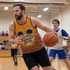 A man wearing a vibrant orange and yellow tank top dribbles a basketball on a gym court, with players in the background.
