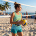 A woman in a light green active tank top holds a volleyball on a beach with palm trees in the background. The top has a logo.