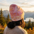A full-colour beanie in shades of pink and orange, worn by a person against a scenic mountain backdrop.