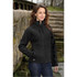 A woman stands by a stone pathway wearing a black soft shell jacket, featuring a zippered pocket and a logo.