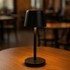 A black mini rechargeable lamp with a tapered shade, standing on a circular base, placed on a wooden table.