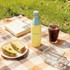 A drink bottle in pale yellow with a light blue cap, set on a picnic blanket with food and a beverage.