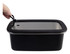 A hand holding the lid of a black rectangular stainless steel container with a clear plastic top.