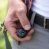 A black carabiner compass thermometer hook is held in a hand, attached to a belt, against a blurred outdoor background.