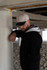 A man leans against a concrete pillar wearing a black jacket and a beige and black cap.