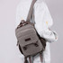 A grey canvas sling backpack hybrid with brown accents, worn on a person's shoulder. It has a logo on the front.